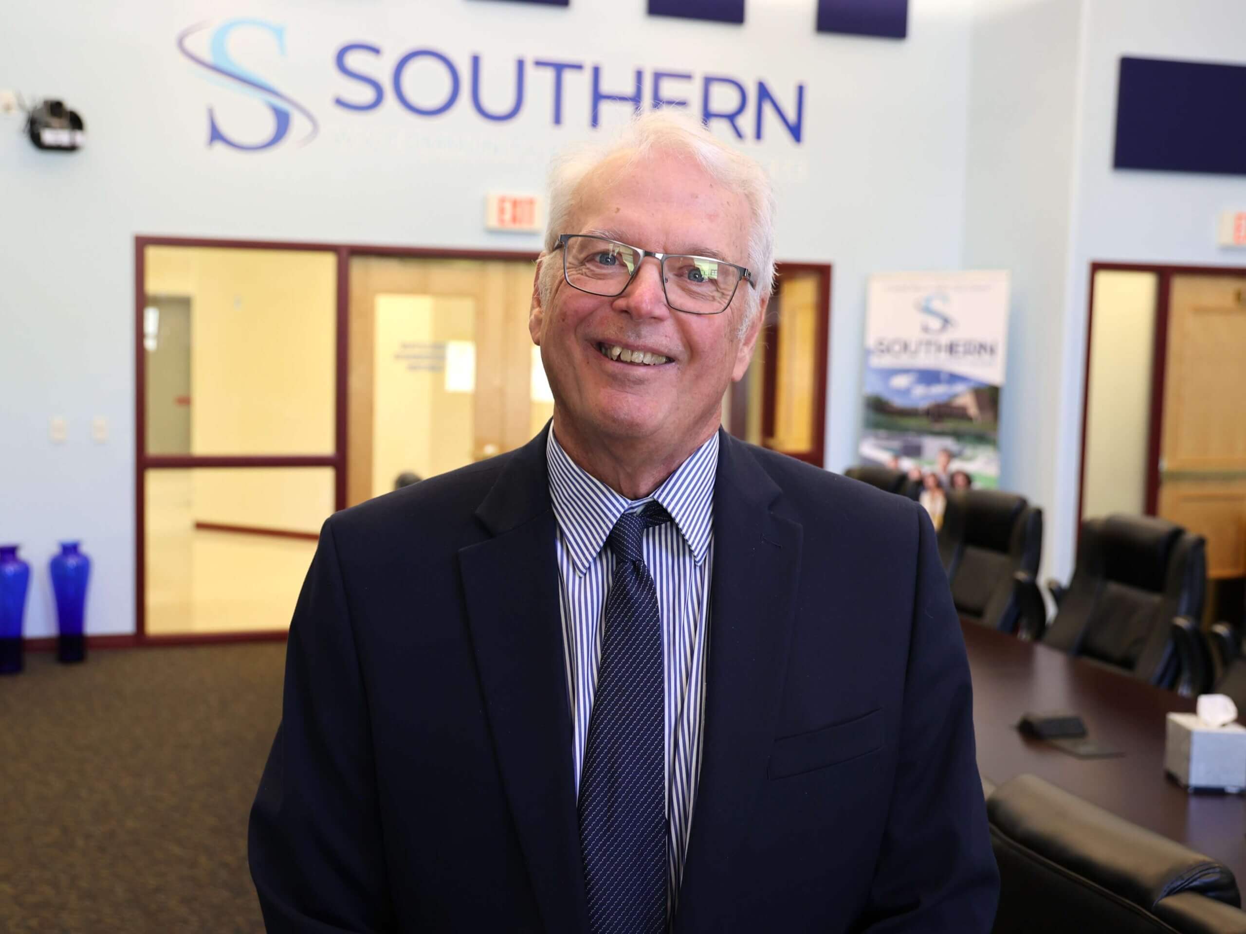 Dr. Corley Dennison, wearing a navy suit and striped tie, stands smiling in a conference room at Southern West Virginia Community and Technical College, with the college’s logo in the background.